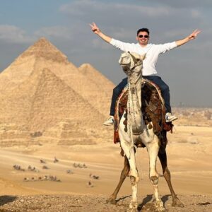 A sandy landscape with the iconic Giza Pyramids and a camel with a rider in the foreground, showing the scale of the monuments